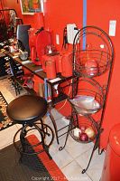 Photo showing metal stool with padded seat adjacent to three-tier wrought iron basket shelf against red wall.