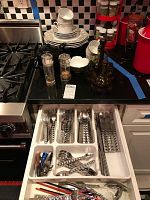 Overview of Fitz and Floyd dinnerware stacked on counter including plates, cups, and saucers near stove, with wooden condiments caddy, glass teapot, salt and pepper shakers nearby. Drawer open below showing stainless steel flatware neatly organized with unique pyramid pattern on handles.