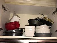 Photo of cabinet shelf showing various bakeware including white ceramic casseroles, metal pans, red silicone bowl or steamer, and wire rack