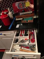 Wide view of kitchen drawer containing various small kitchen tools including can openers, peelers, graters, hand juicers, pizza cutters with red, white, and black handles.