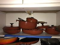 Eight vintage Le Creuset pots and pans stacked on a shelf, showing their orange-red enamel finish and wooden handles.