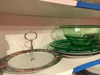 Porcelain and green glass platters stacked on white shelf. Porcelain platter with jungle animal border visible and green glass oval spatter on top of two black and white patterned plates.