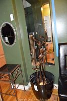 Full view of tall metal fountain sculpture in black plastic pot, showing curved drip stalks and base setup in a room corner with mirror and furniture.