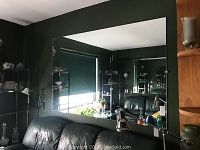 Mirror shown mounted on dark green wall reflecting living room including black leather couch, shelves with glassware, and lamp.