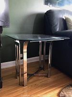 Full view of the glass topped side table placed next to black leather furniture, showing its rectangular shape and reflective metal legs.