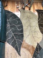 Rug showing overlapping large leaf shapes in green, beige, brown, and cream colors alongside hardwood floor and part of furniture.