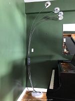Full view of tall metal floor lamp with five curved arms ending in chrome reflective lampshades next to a piano.