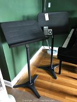 Two black metal music stands with tripod bases, placed near a piano against a green wall. One stand has a Seiko Quartz metronome resting on its platform.