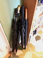 Five black umbrellas standing against a wall, showing various handle styles including curved wooden and textured wooden handles.