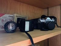 Full view of Canon AE-1 camera with attached lens, extra lens, flash unit, manuals and case on shelf