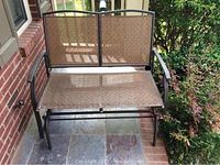 Front view of two-person outdoor glider showing woven backrest and seat, metal armrests and frame, placed on tiled porch.