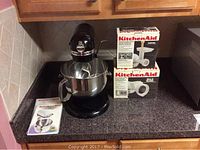 Front view of mixer on countertop with whisk in bowl, manuals and two boxed attachments stacked beside it
