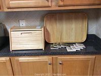 Light brown oak bread box and Studio Nova light brown wooden tray side by side on kitchen counter with eight coasters in front of tray
