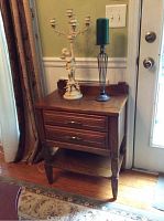 Dark brown wooden side table showing two drawers with handles and bottom shelf. On top are a tall dark pewter colored candle holder with green pillar candle and an ivory cherub candelabra with several small cherubs and multiple candle holders.
