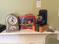 Photo of the Kodal Vigilant camera with box, Howdy Doody metal lunch box, and Weill Disney Mickey Mouse clock on a white shelf.