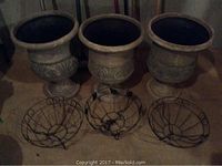 Three large resin garden pots with ornate pattern along the center and pedestal, placed on floor alongside three black circular wire hanging baskets.