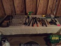 Wide view of folding table covered with a white cloth holding garden tools, fishing rods with reels, and other miscellaneous items in a basement setting.