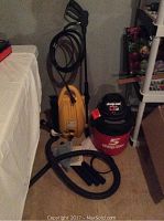 Full lot photo showing yellow electric power washer with black hose and wand next to a red and black Shop-Vac on carpeted floor in basement.