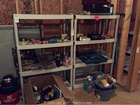 Two white plastic shelving units shown against a plywood wall in a basement, loaded with various tools and items.