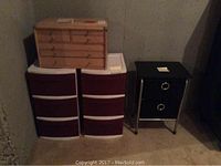 Five storage units grouped in basement corner: two plastic maroon-white three-drawer units, one wooden multi-drawer unit, one small black two-drawer unit with silver frame on right.