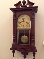 Front angled view of entire wooden Daniel Dakota wall clock showing carved details and pendulum behind glass panel.