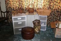 Two wicker baskets on top of an off-white round decorator table with a mirrored top, flanked by two Sterilite plastic drawer carts with three drawers each, one with wheels and one without. Brown round ottoman and decorative lion container placed in front.
