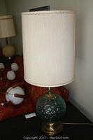 Side view of aqua textured glass ball base table lamp with cylindrical cream fabric shade and brass base.