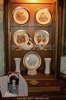 Display cabinet with Lenox Boehm decorative plates showing detailed woodland animal artwork, a white porcelain swan, two white Gorham vases, a floral porcelain vase, and a single decorated plate on stand, plus a collectible doll in a box folded out on lower shelf.