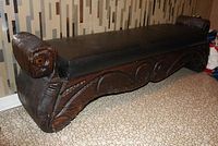 Full view of the carved wooden bench showing the black padded seat and carved scroll detail on legs and frame.