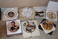 Overview photo showing seven collectible decorative plates including the Andrew Wyeth plate The Kuerner Farm, Hummel plate from 1974, and other assorted plates with various artistic and nostalgic themes.