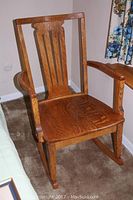 Full view of oak rocking chair with curved armrests, upright slatted back, and wooden rockers on carpeted floor.