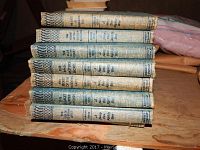 Stack of vintage hardcover books with blue-green patterned spines featuring Nancy Drew, Judy Bolton, Beverly Gray titles, pink chenille bedspread and metal trays stacked behind.