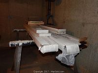 Boxes of flooring stacked on sawhorses in basement. Four long boxes mostly wrapped in white packaging, a few shorter boxes on top with visible wood finish ends.
