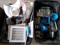 Photo of plastic tub and bin with assorted electrical and plumbing supplies. Items include white PVC plumbing fittings, multiple electrical outlet boxes in blue plastic, white vent covers, and electrical outlets.
