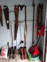 Photo showing hanging vintage hand saws with wooden handles along with bar clamp, hand rakes, and leaf grabber on wall with boots and Toro snow shovel on floor.