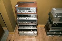 Stack of six vintage amplifier receivers featuring wooden and metal casing, varying sizes and configurations, photographed from the front showing knobs, sliders, and analog displays.