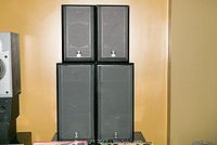 Four BMW speakers in black wooden cabinets with gray speaker grille cloth, stacked in pairs. BMW logos visible on speaker cloth. One cabinet has rear damage present.