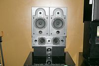 Front view of two Energy model 2.1e speakers stacked above two Energy centre speakers on a black surface against a brown wall. All speakers missing grills.