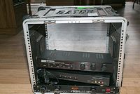 Overall view of Gator rack mount case with three electronic devices (Hughes Retriever, JVC AV selector, Gemini PL-7) stacked inside on a hardwood floor in a room with wood furniture.