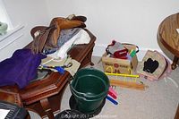 Wide view showing Carlyle saddle on wooden table with purple blanket, green bucket, grooming brushes, riding crops, and box with accessories on carpet