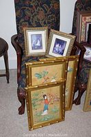 Three Oriental style pictures in gold bamboo-style frames stacked on a chair, two smaller framed prints of children placed behind them on another chair.