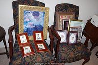 Overall view of two patterned fabric chairs with various framed artwork arranged on and behind them including a large gold-framed painting of a woman, smaller wooden framed portraits, butterfly and floral prints, and street scene.