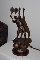 Bronze-tone tennis player figurine with racket raised, standing on detailed base with tennis balls and rackets.