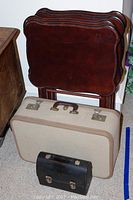 Four wood TV tray tables stacked, beige hard-sided suitcase and small black metal lunch box in front.