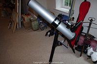 Full view of silver Meade telescope mounted on tripod stand in room, with red carrying case and other household items in background