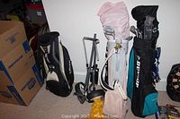 Three golf bags along a wall including a black Callaway, white with pink cover, and Dunlop bags, plus a silver foldable golf push cart and bag of yellow golf balls.