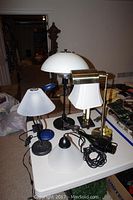 Photo showing 7 lamps on a table with various shades and bases, some glass, some metal, cords visible.