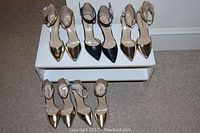 Five pairs of new women's heels displayed on carpet and white box, mixed gold metallic and black all with ankle straps.