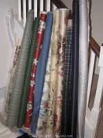 Photo showing eleven bolts of fabric standing upright next to a wall and a staircase railing in the background. Fabrics have various colors and patterns including floral, plaid, geometric, and solids.