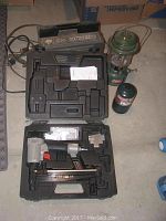 Photo showing Porter-Cable nail gun in black plastic case, Coleman green lantern and green fuel container on concrete floor.